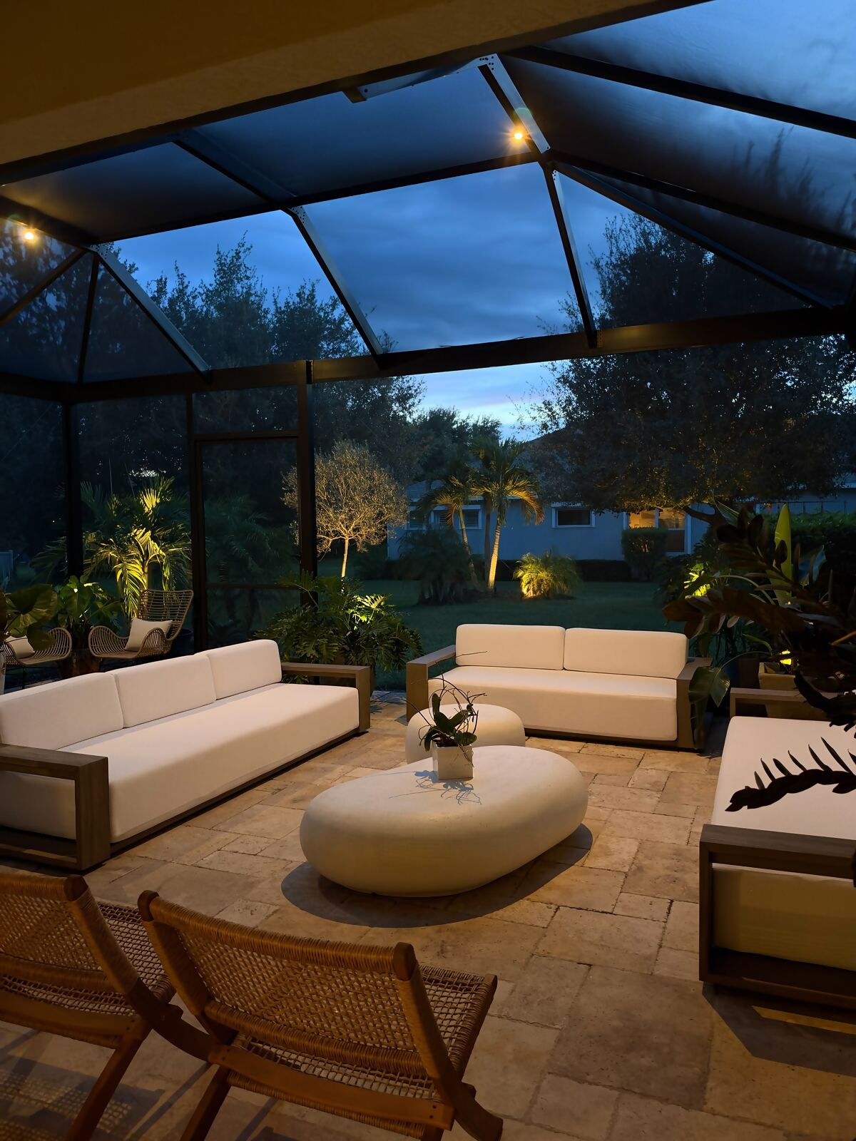 Outdoor seating area inside a screened lanai, softly lit at dusk with uplit plants beyond