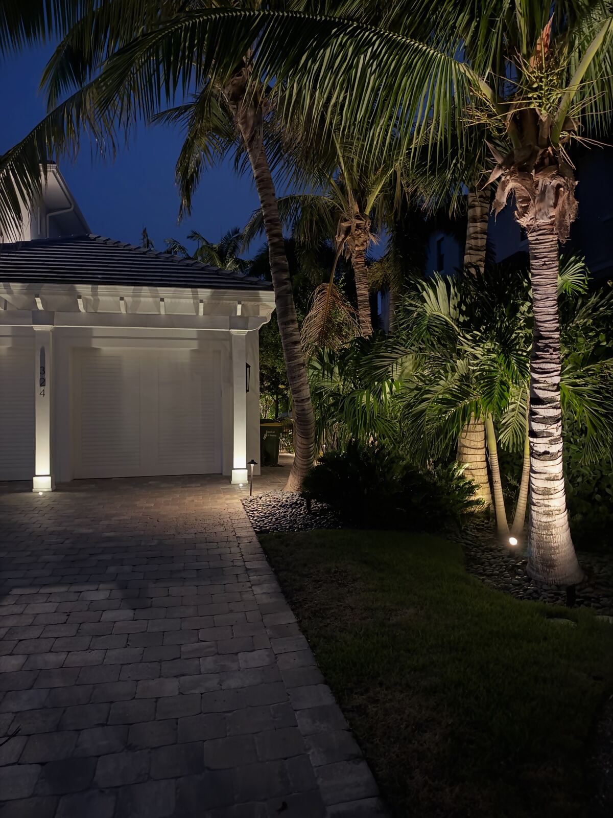 Paver driveway and garage framed by uplit palms and garden lighting at night